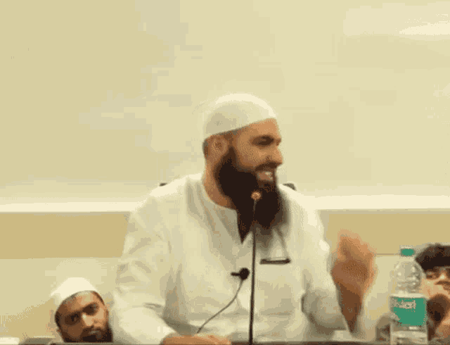 a man with a beard is giving a speech in front of a microphone with a bottle of aquafina water in the background