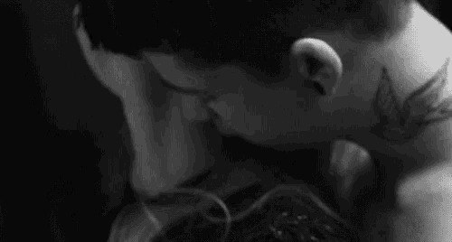 a man and a woman are kissing in a black and white photo . the man has a tattoo on his neck of a bird .