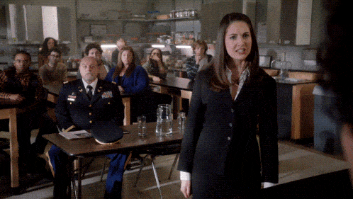 a woman in a black suit stands in front of a group of people including a man in a military uniform