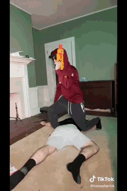 a man in a red hoodie is kneeling on another man holding a nerf gun