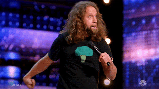 a man with long hair and a broccoli shirt is holding a microphone on a stage