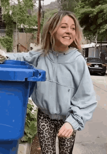 a woman wearing a blue jacket and leopard print leggings is standing next to a blue trash can and smiling
