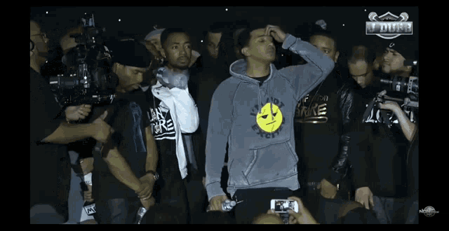 a man wearing a grey hoodie with a smiley face on it stands in front of a crowd