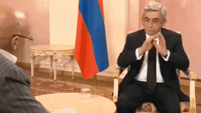 a man in a suit and tie is sitting at a table talking to another man in front of a flag .