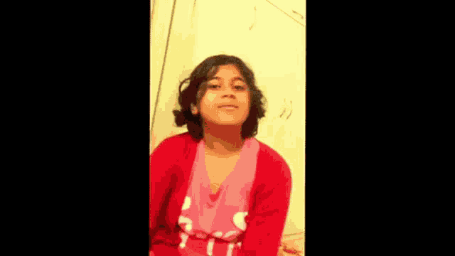 a young girl wearing a red sweater and a pink shirt is sitting down and smiling .