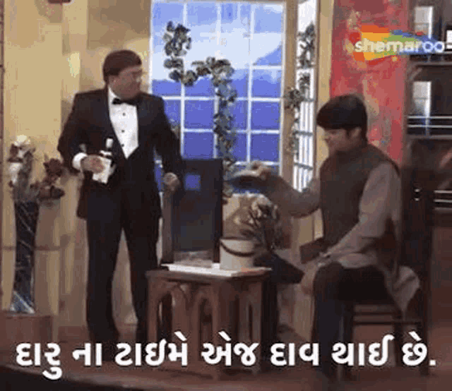a man in a tuxedo is standing next to a man sitting on a chair in a room .