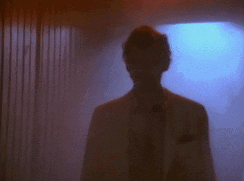 a man in a white suit and tie is standing in a dark room with a blue light behind him .