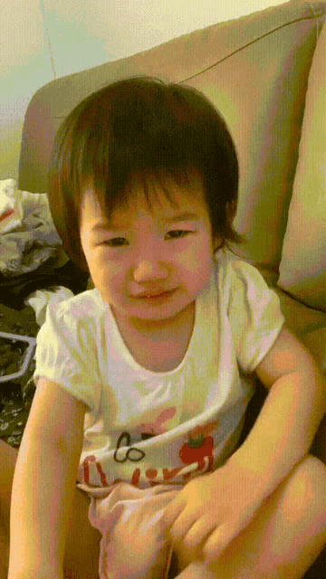 a little girl is sitting on a couch wearing a white shirt with a strawberry on it