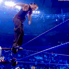 a wrestler in a black tank top is jumping over ropes
