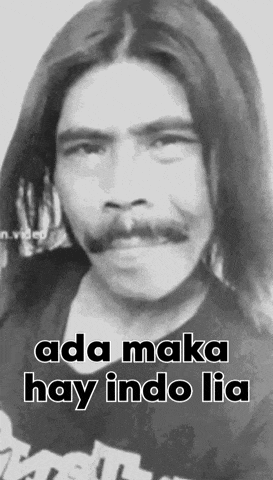 a man with long hair and a mustache is wearing a black shirt with the words ada maka hay indo lia written on it .