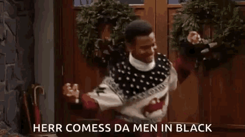 a man in a sweater is dancing in front of a door with the words herr comess da men in black written on it .