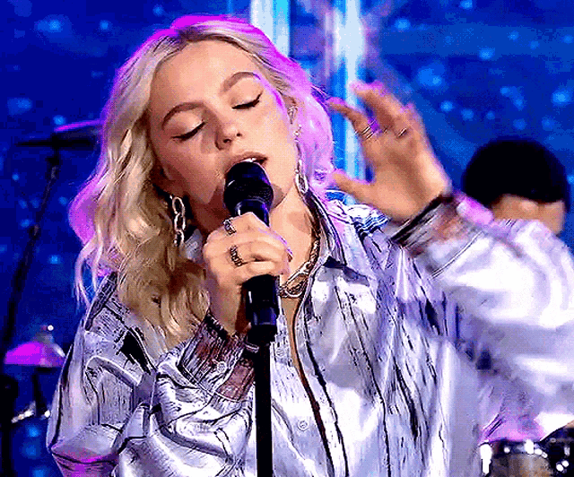 a woman singing into a microphone wearing a silver shirt and earrings