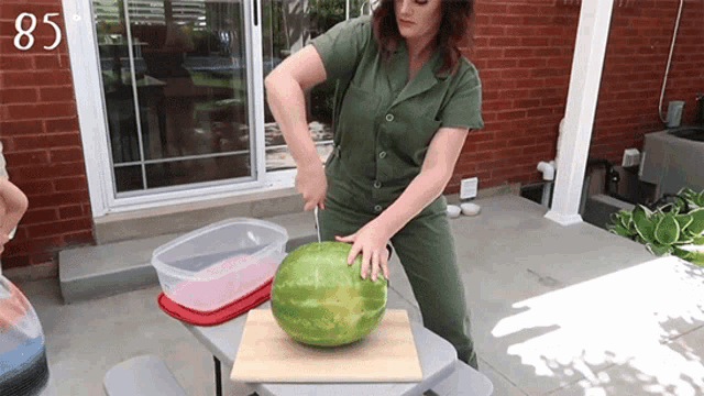 a woman is cutting a watermelon with a knife on a table with the number 85 on the wall behind her