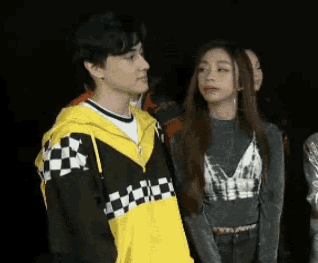 a man in a yellow and black checkered jacket stands next to a woman in a gray top