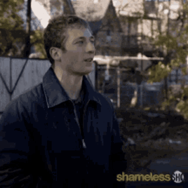 a man in a blue jacket is standing in front of a fence with the words shameless written on the bottom