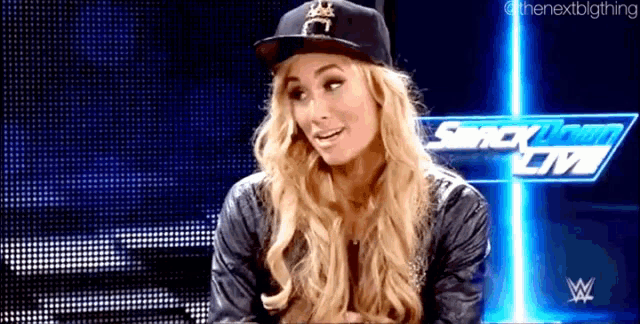 a woman wearing a baseball cap and a leather jacket is sitting in front of a screen that says smack down on it