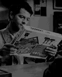 a black and white photo of a man sitting at a table reading a newspaper .