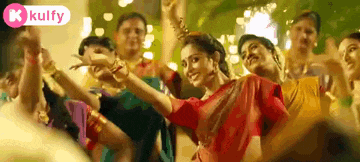 a woman in a red saree is dancing in front of a crowd with a kolfy logo in the background