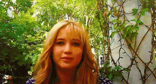 a woman with long blonde hair is standing in front of a lush green forest .