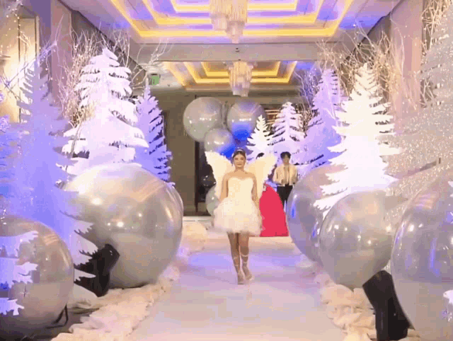 a woman in a white dress is walking down a runway surrounded by balloons and trees