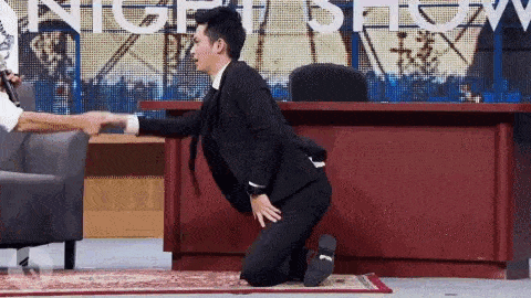 a man in a suit is kneeling down in front of a desk while a woman shakes his hand .