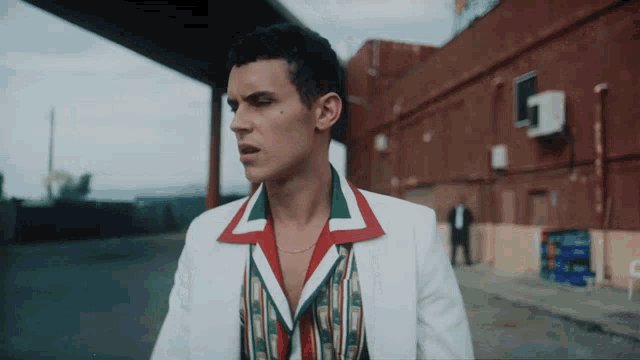 a man in a white suit with a red and green collar stands in front of a building with the letter g on it