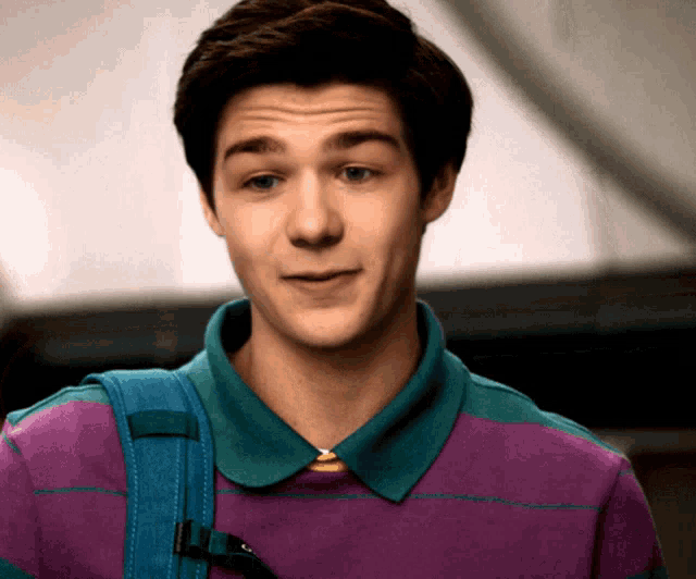 a young man wearing a purple shirt and a blue backpack looks at the camera