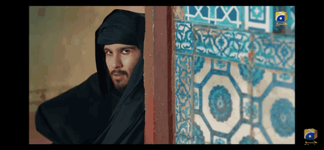a man in a black turban stands in front of a wall with blue and white tiles