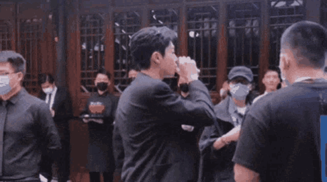 a man in a suit is drinking from a cup while standing in front of a crowd of people wearing masks .
