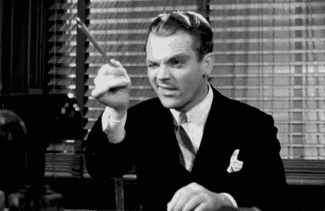 a man in a suit and tie is sitting at a desk holding a pen and pointing at the camera .