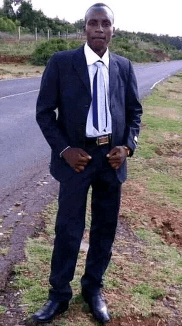 a man in a suit and tie is standing in the grass next to a road .