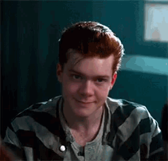 a young man with red hair is wearing a striped shirt and smiling in a jail cell .