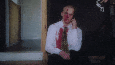 a man with blood on his shirt and tie is talking on a cell phone