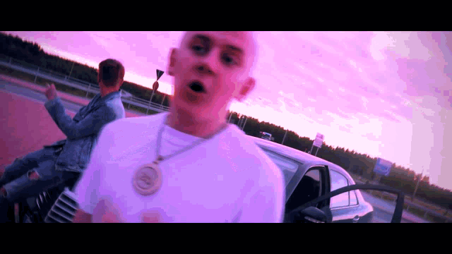 a man wearing a necklace with the letter c on it stands next to a car