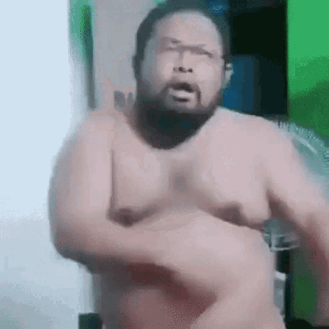 a shirtless man with a beard is standing in front of a fan in a room .