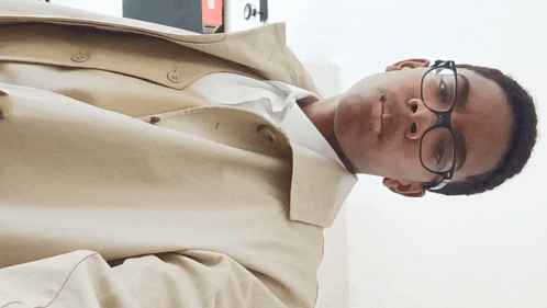 a young man wearing glasses and a trench coat looks at the camera