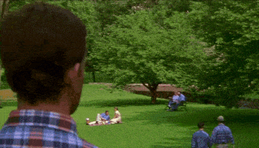 a man in a plaid shirt looks at a group of people having a picnic in a park