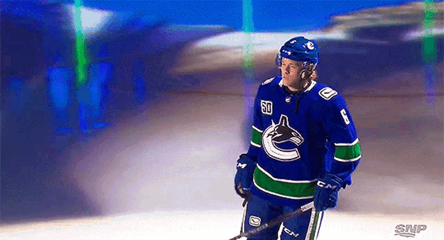 a hockey player in a blue and green uniform is standing on the ice holding a stick .