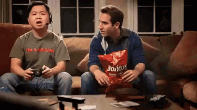 two men are sitting on a couch playing video games and one of them is holding a bag of doritos