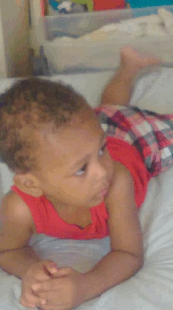 a baby in a red tank top and plaid shorts is laying on his stomach on a bed