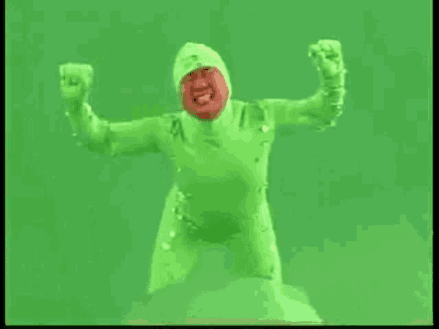 a man in a green suit is standing in front of a green screen with his arms in the air .