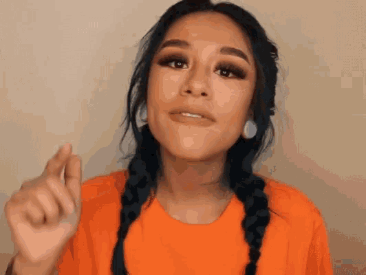 a woman with braids and earrings is wearing an orange shirt and making a funny face .