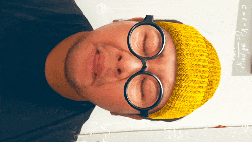 a man wearing glasses and a yellow hat with mathematical equations on the wall in the background