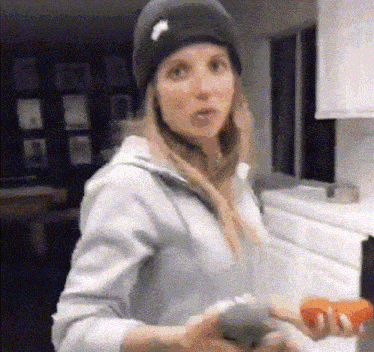 a woman wearing a beanie and a hoodie is standing in a kitchen holding tomatoes .
