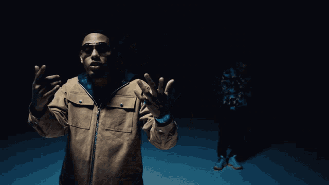 a man in a brown jacket and sunglasses stands in a dark room with his hands outstretched