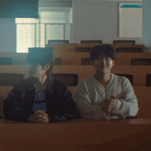 two young men are sitting in a classroom with a sign on the wall that says " exit "