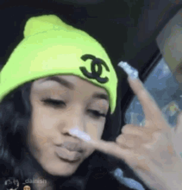 a girl wearing a neon yellow beanie with a chanel logo on it is making a peace sign .