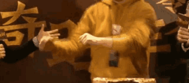 a man in a yellow hoodie is standing in front of a cake and making a hand gesture .