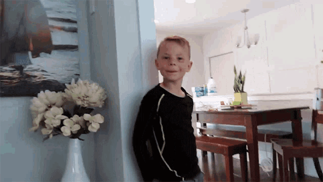 a young boy in a black shirt is standing next to a table with flowers in a vase