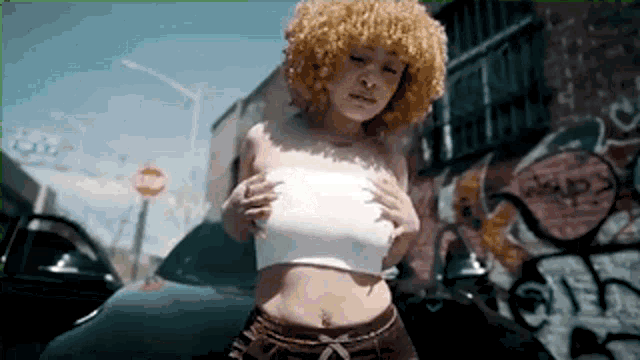 a woman with curly hair is standing next to a car in front of a graffiti covered wall .